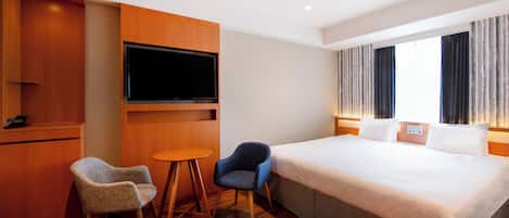A hotel room with a large bed, a television mounted on the wall, two chairs, a small table, and a window with curtains.