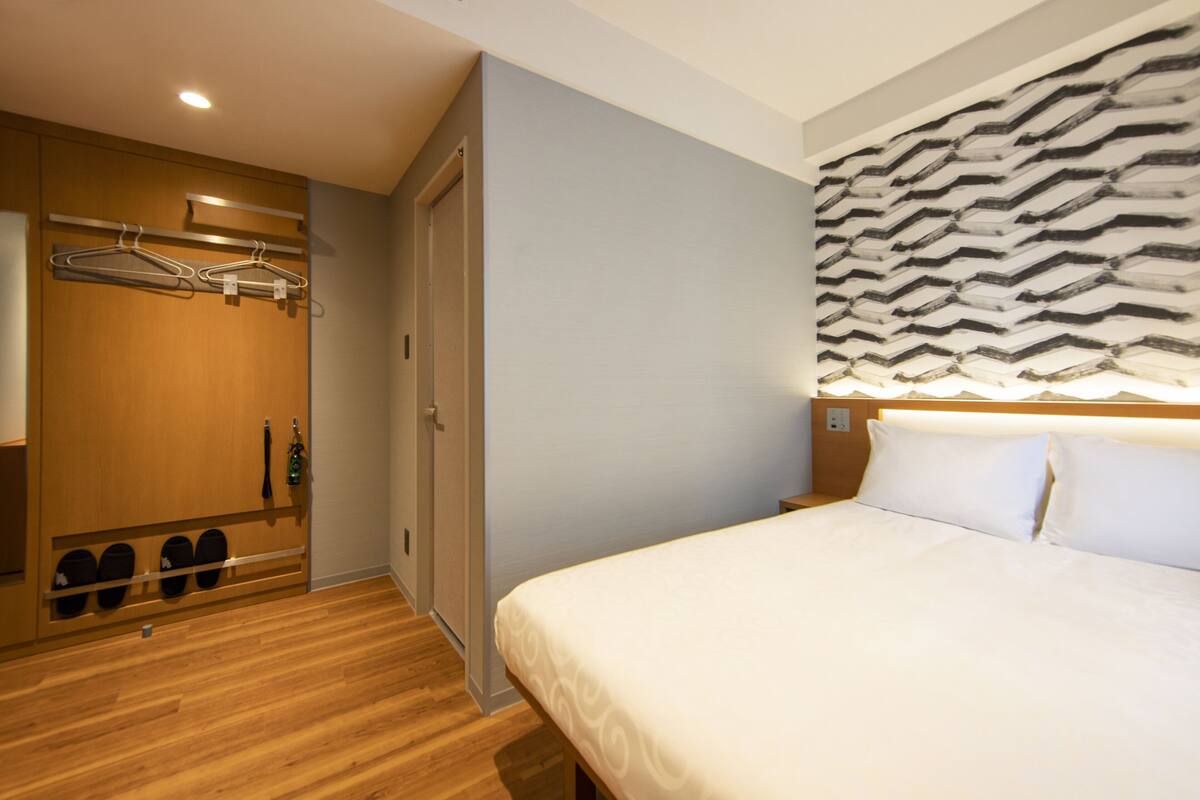 A hotel room with a bed, a wardrobe with hanging clothes, and a shoe rack.