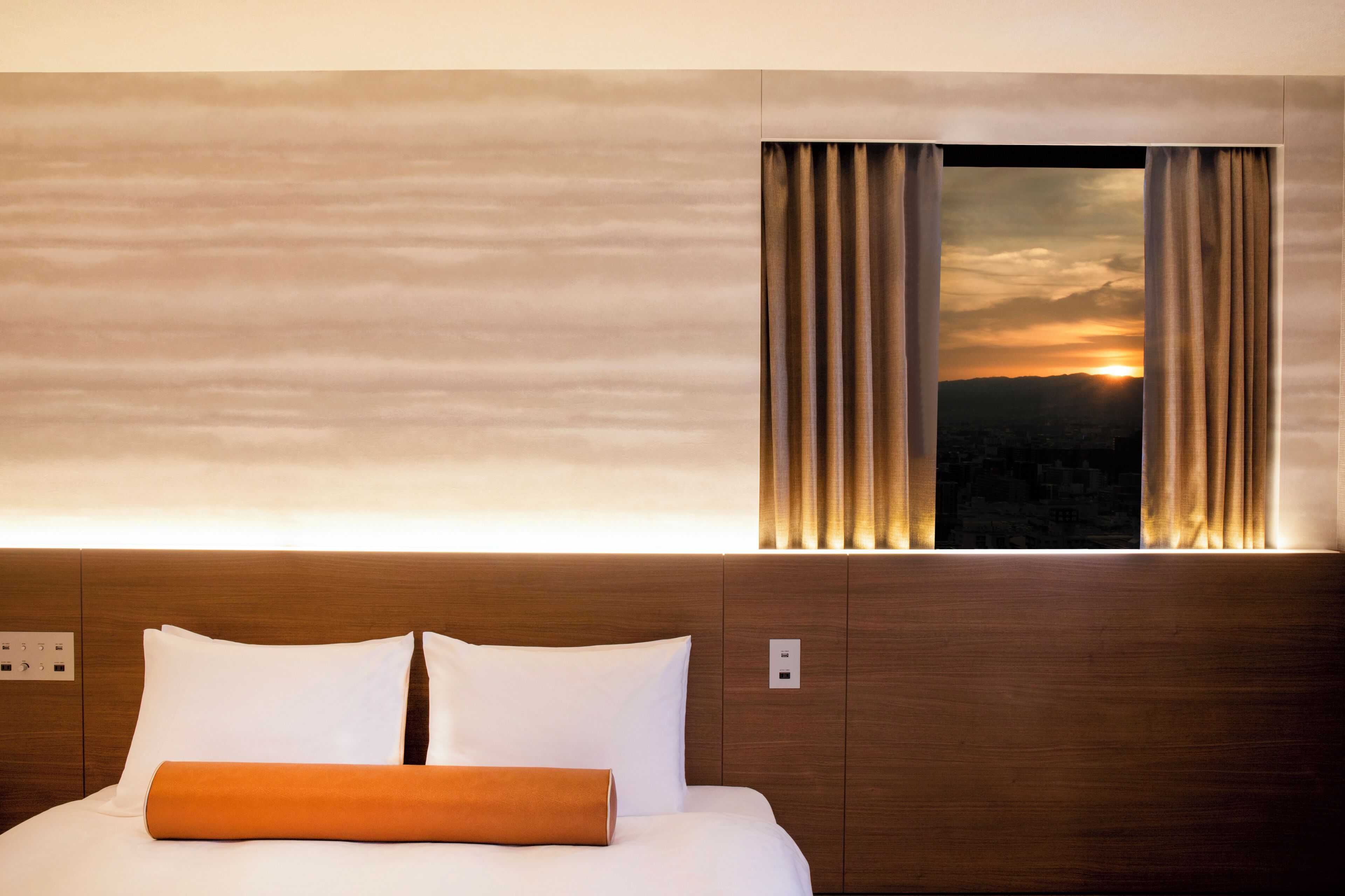 A hotel room with a bed, a nightstand, a window with curtains, and a view of the sunset.