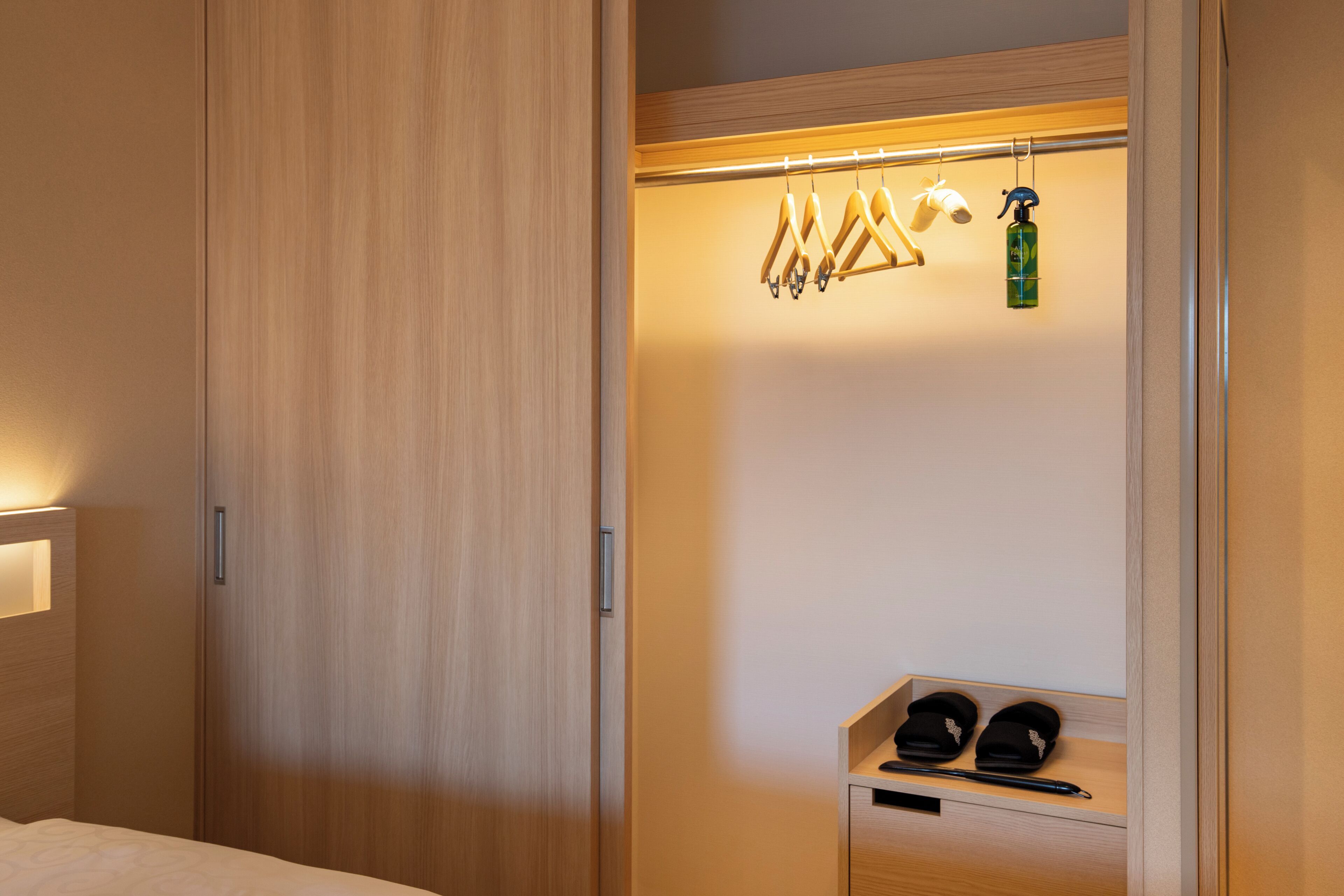 Habitación de hotel con armario de madera, perchero, estante con pantuflas y un bolígrafo, y una botella de gel hidroalcohólico.