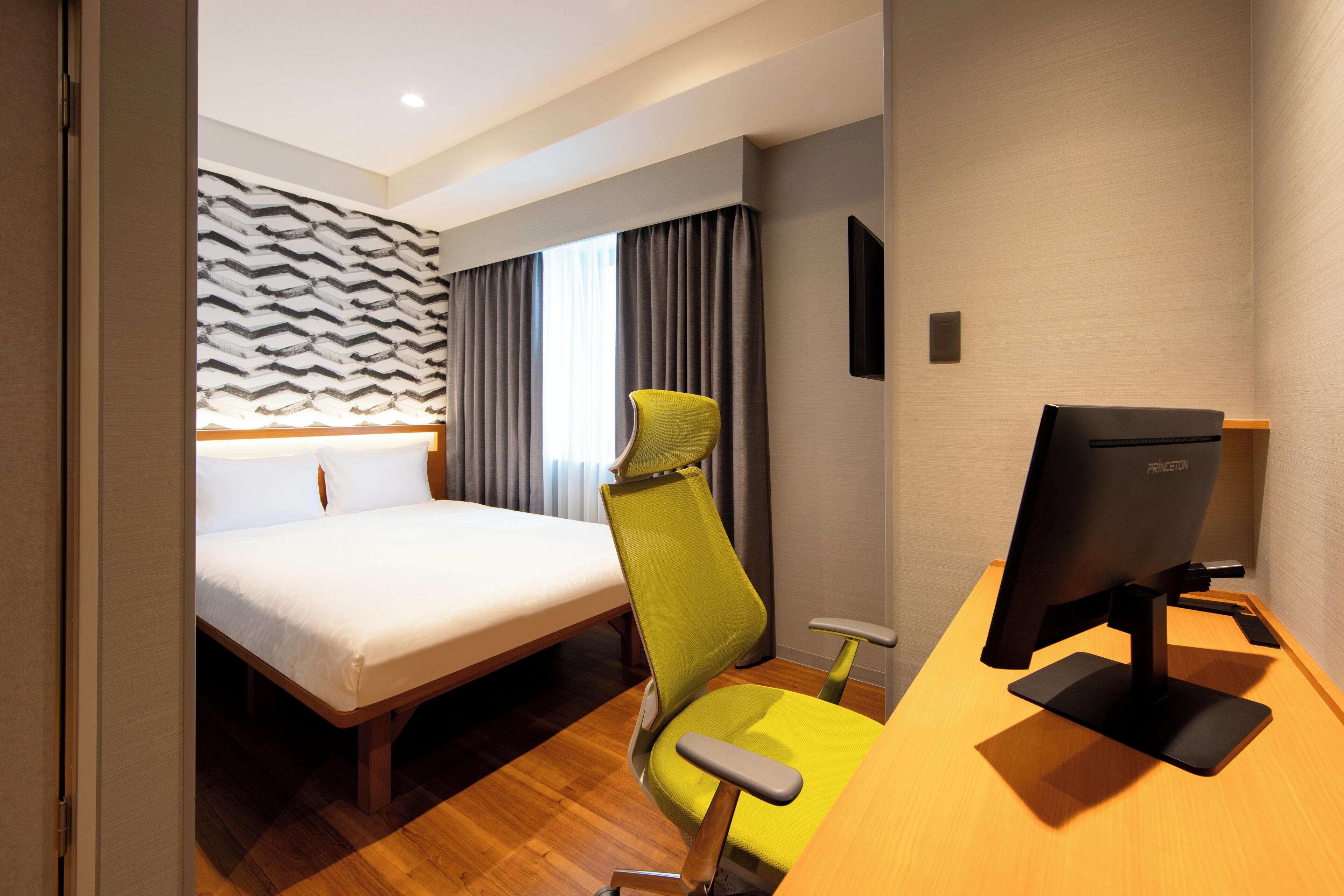 A hotel room with a bed, a desk with a computer, a chair, and a window with curtains.