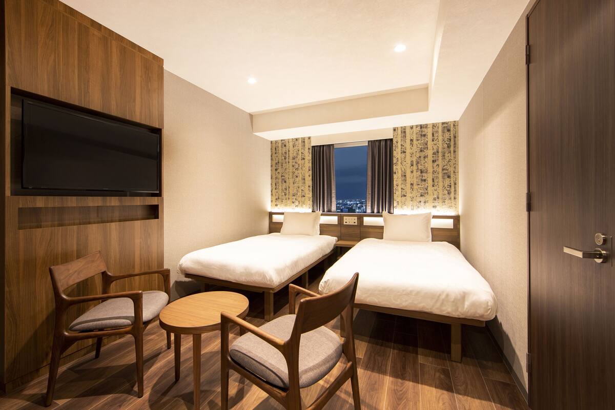 A hotel room with two single beds, a TV mounted on the wall, a wooden desk, and a chair.