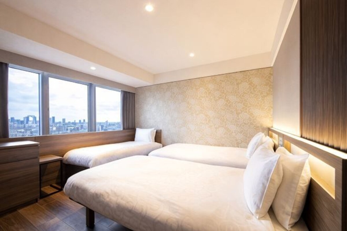 Habitación de hotel con dos camas, un aparador de madera, una mesita de noche y un ventanal con vistas a la ciudad.