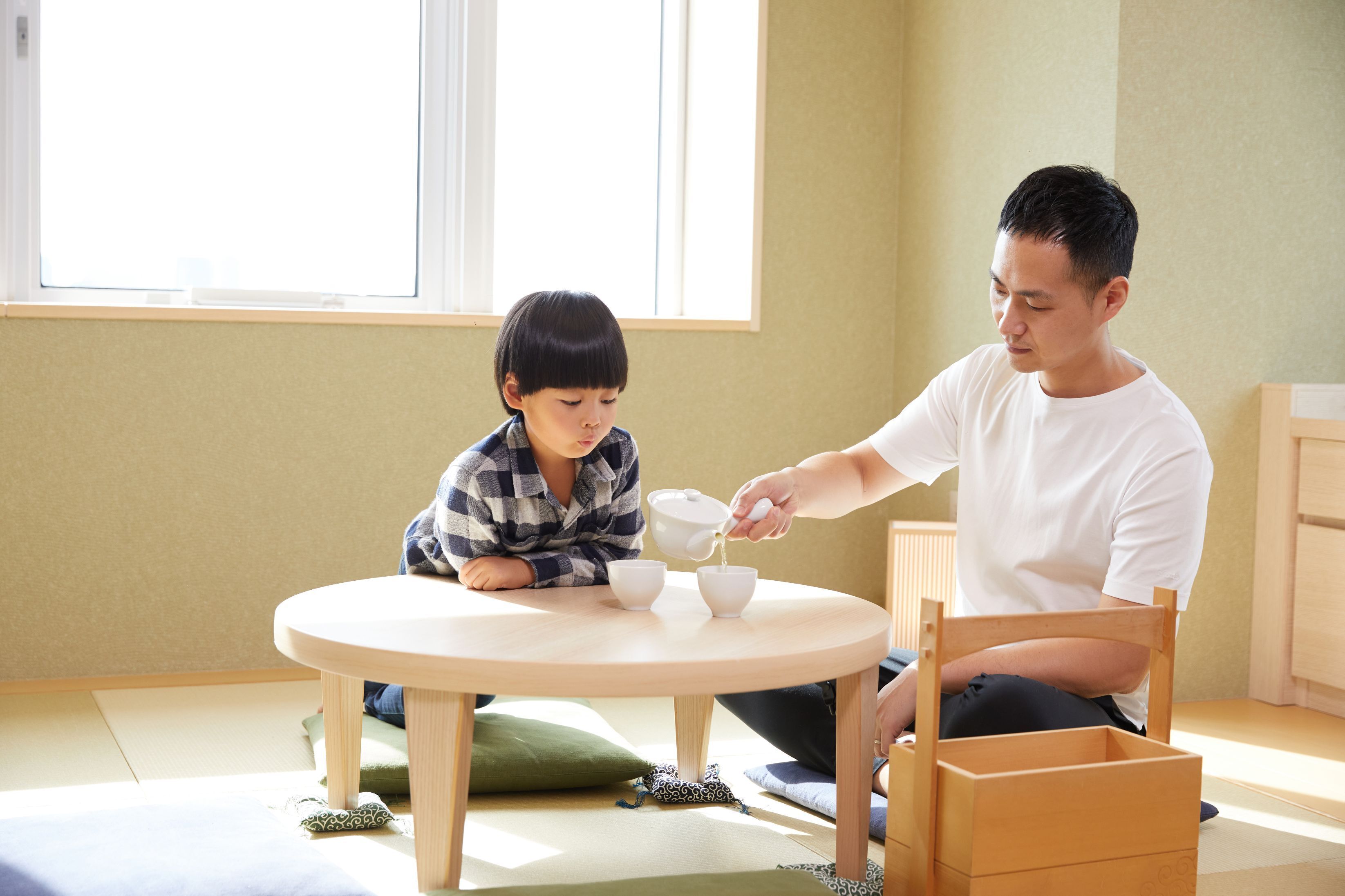 A room with a round table, a child and an adult sitting at it, and a window in the background.