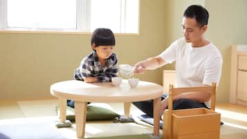 A room with a round table, a child and an adult sitting at it, and a window in the background.