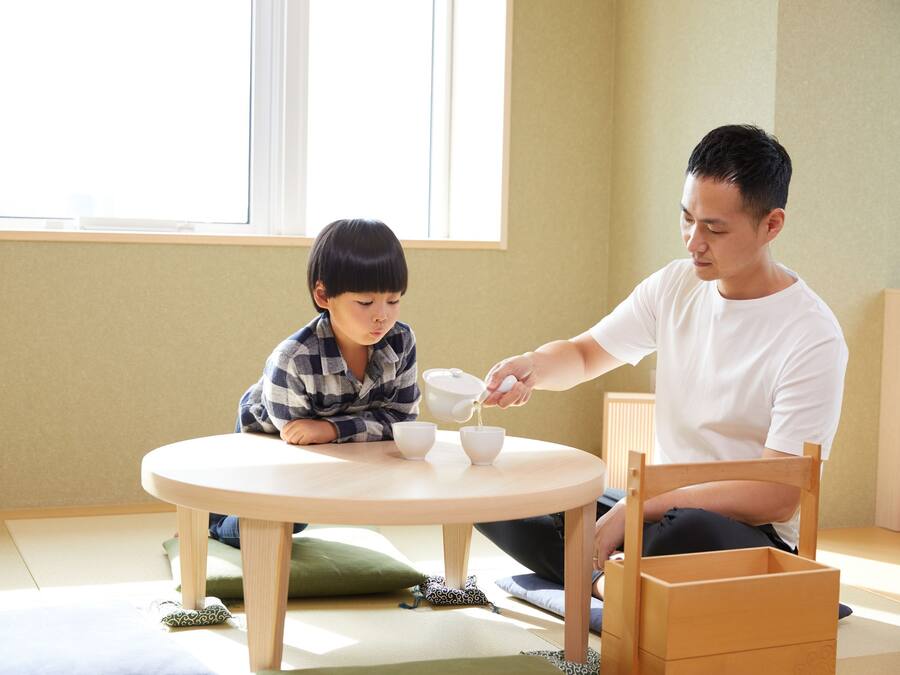 A room with a round table, a child and an adult sitting at it, and a window in the background.