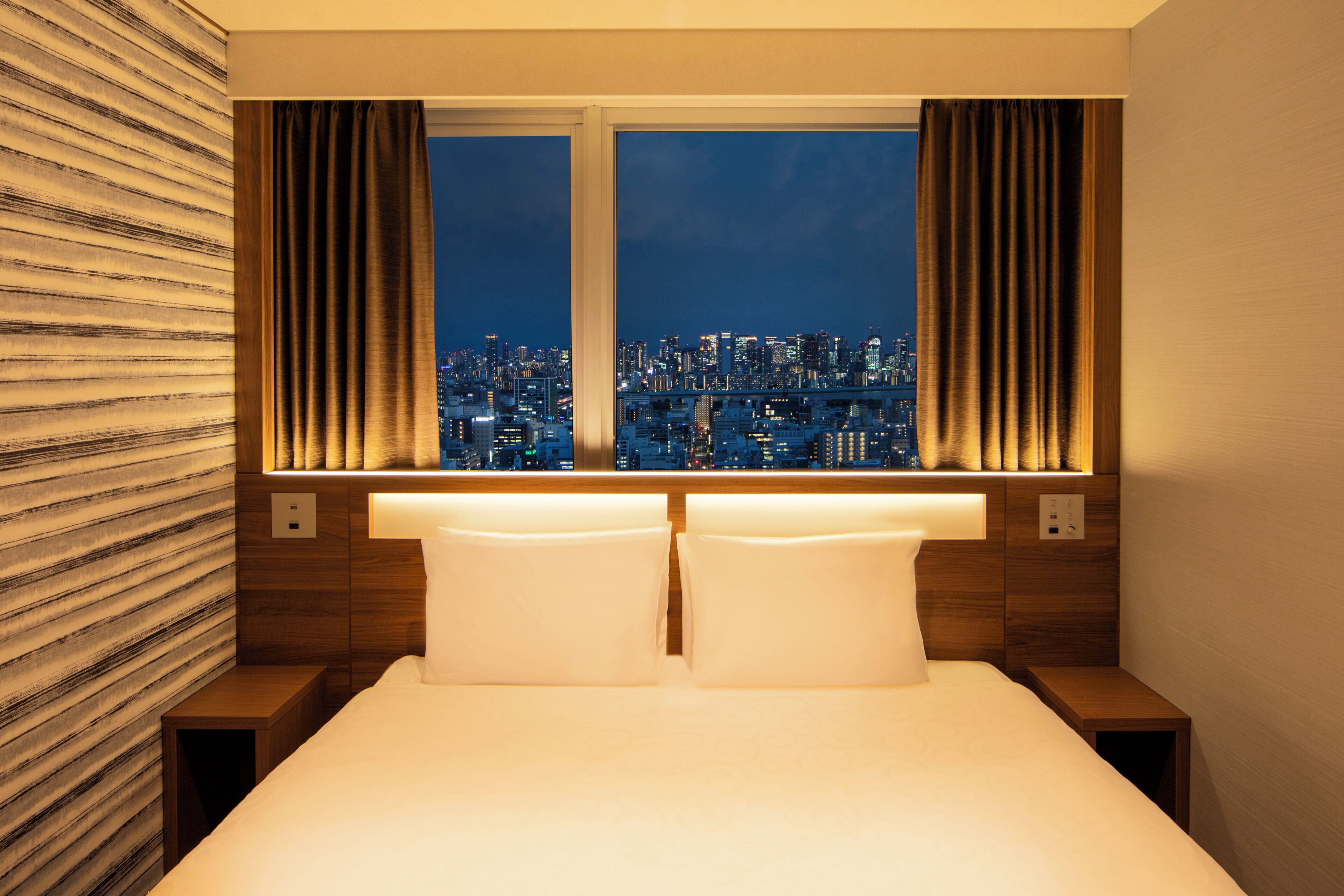 Habitación de hotel con cama, dos mesitas de noche, ventana con cortinas y vista a la ciudad.