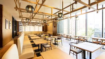 A modern restaurant with long communal tables and booths, large windows offering city views, and a wooden ceiling with hanging spherical light fixtures.