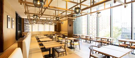 A modern restaurant with long communal tables and booths, large windows offering city views, and a wooden ceiling with hanging spherical light fixtures.