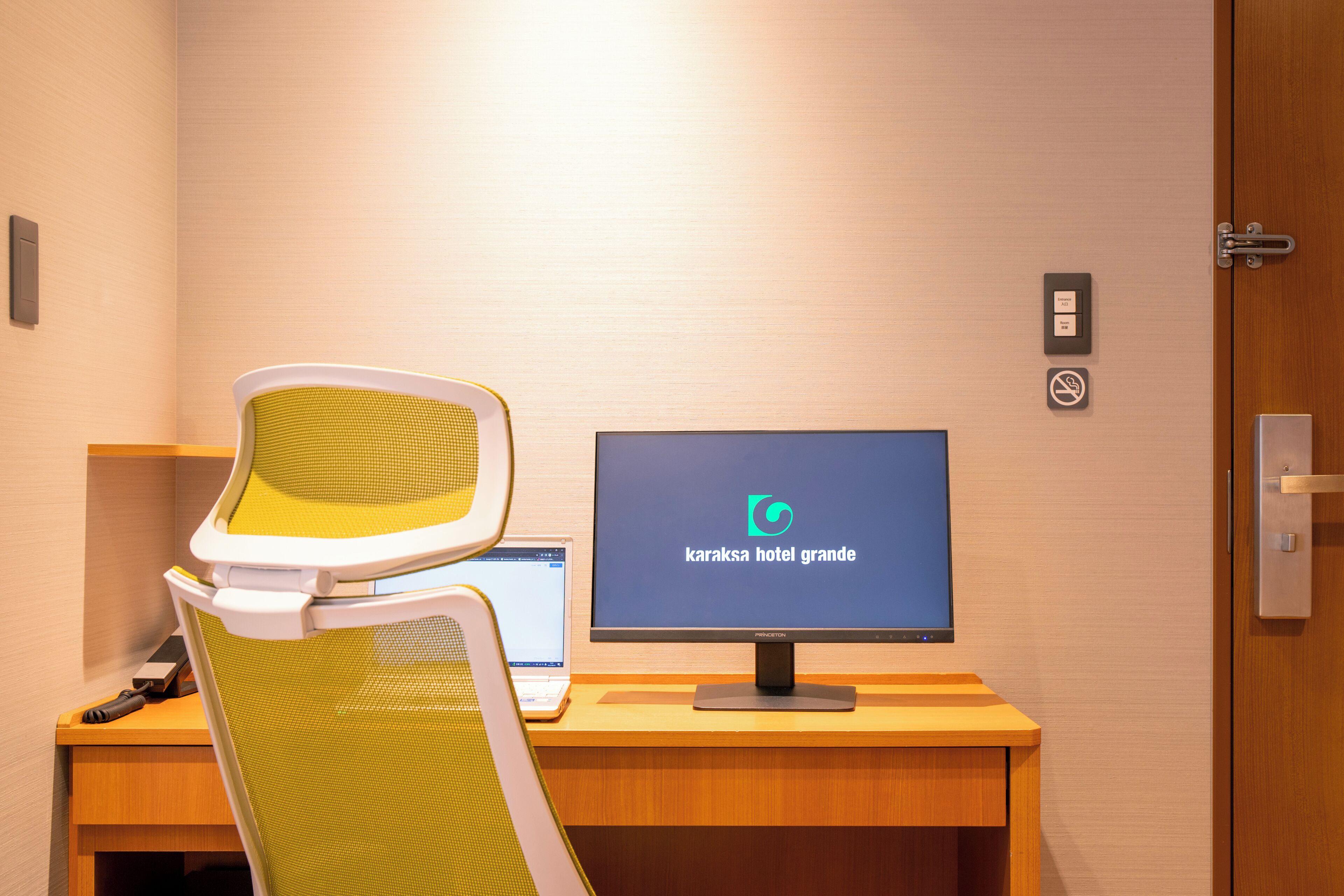 A hotel reception desk with a computer monitor displaying the hotel's name, a chair, a desk, and a door with a lock.