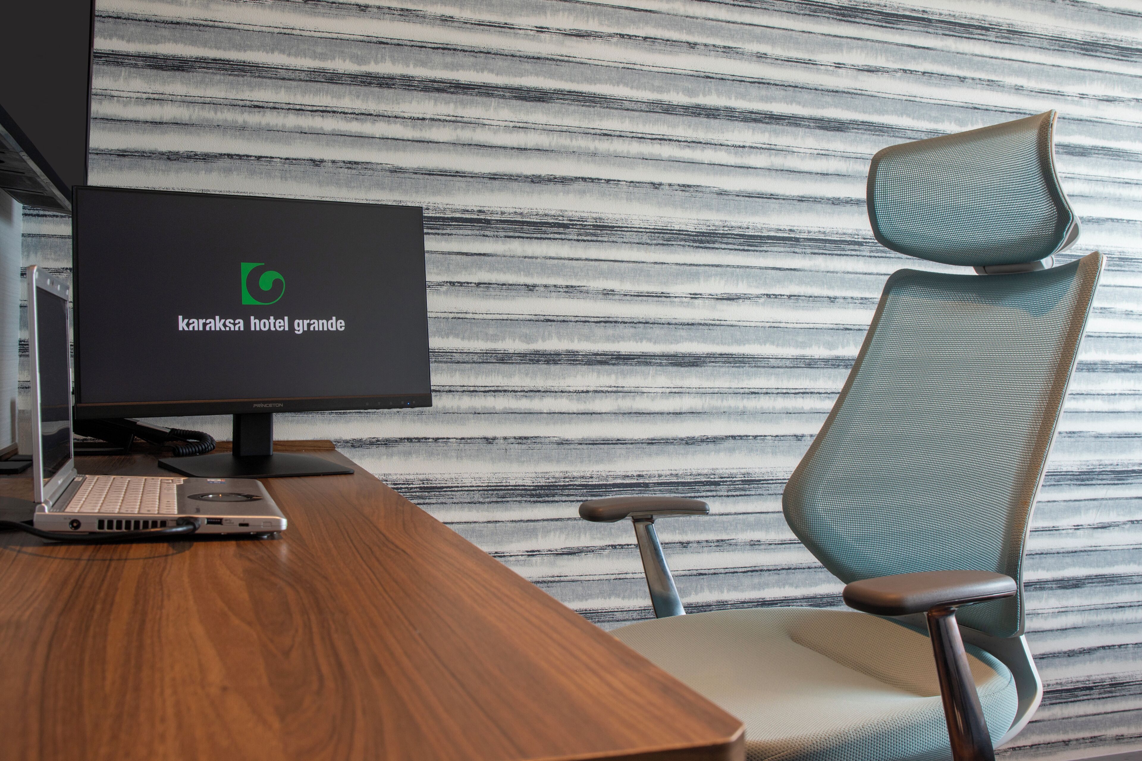 Un espace de bureau moderne équipé d’un ordinateur portable, d’un écran d’ordinateur de bureau affichant le logo d’un hôtel et d’un fauteuil à haut dossier.