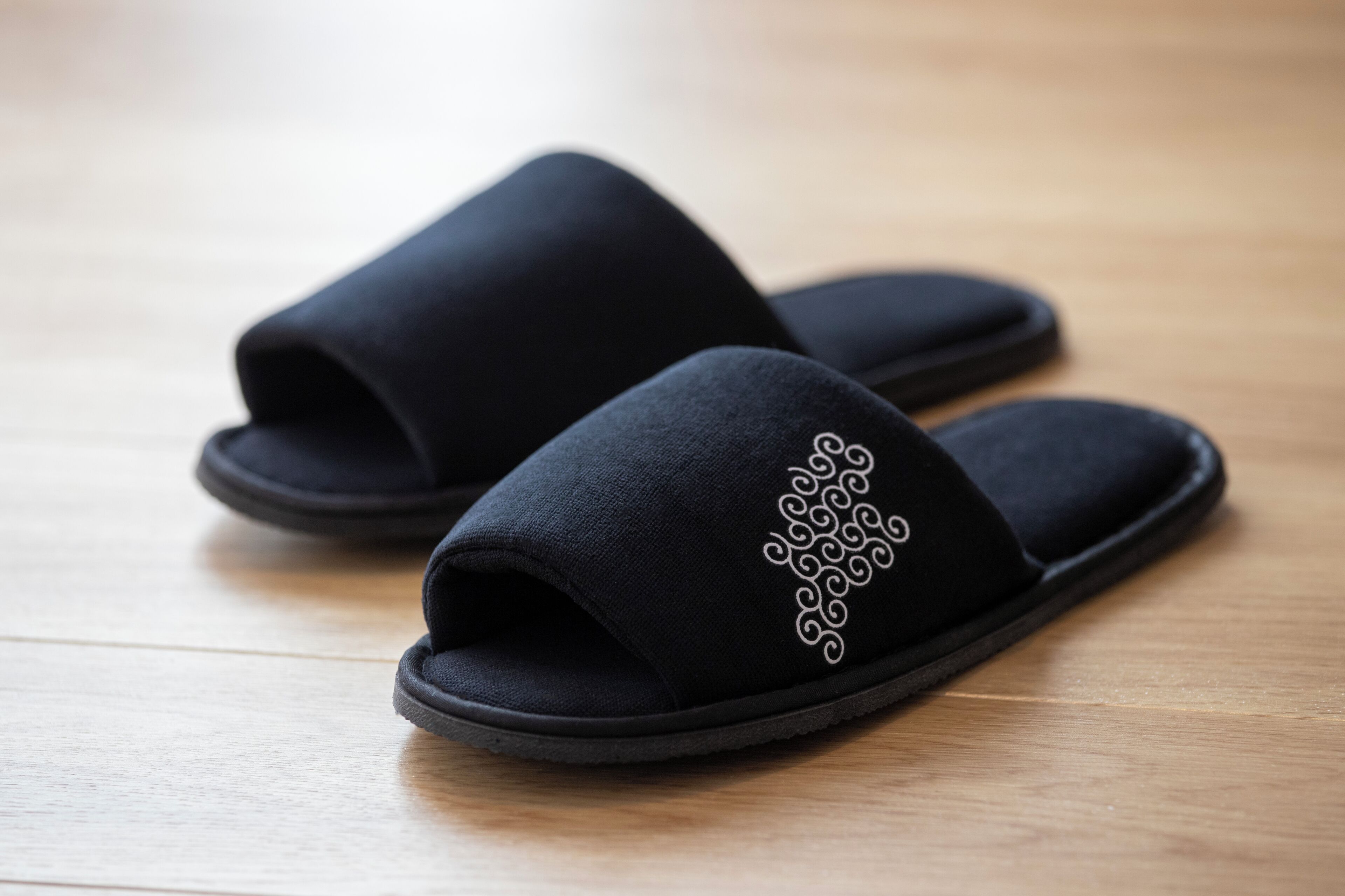 A pair of black slippers with a decorative pattern on the side, placed on a wooden floor.
