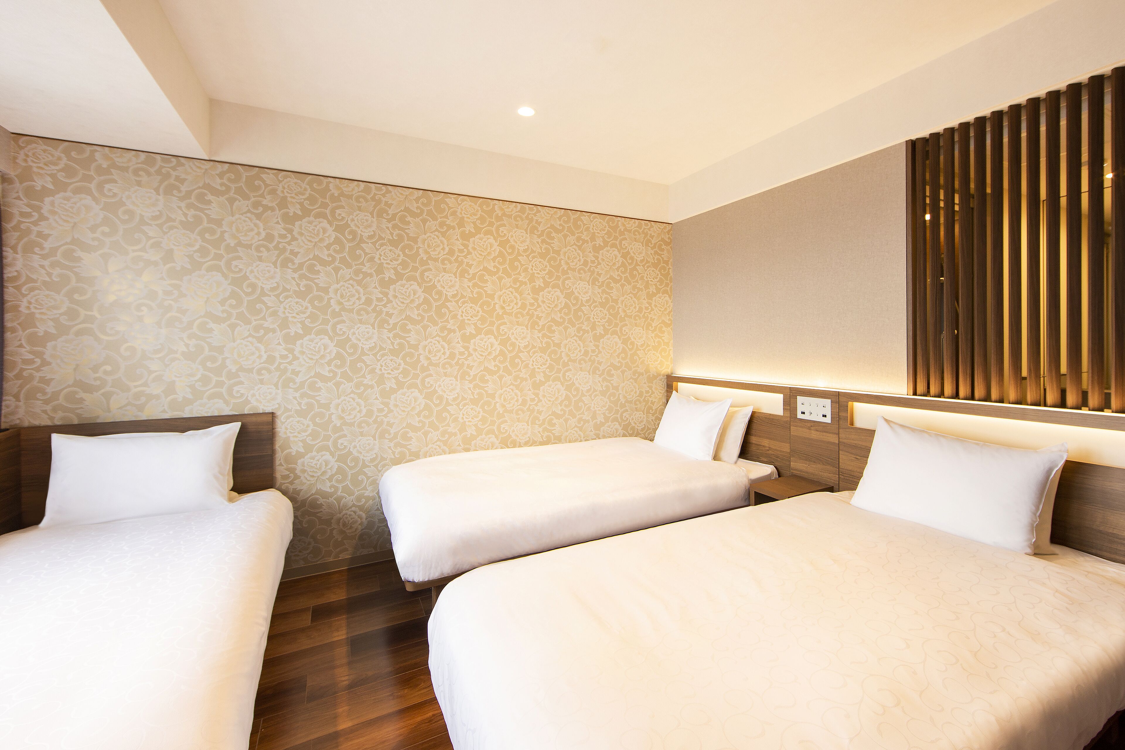 Quarto de hotel com duas camas de solteiro, cabeceira de madeira, papel de parede estampado e luminária moderna.
