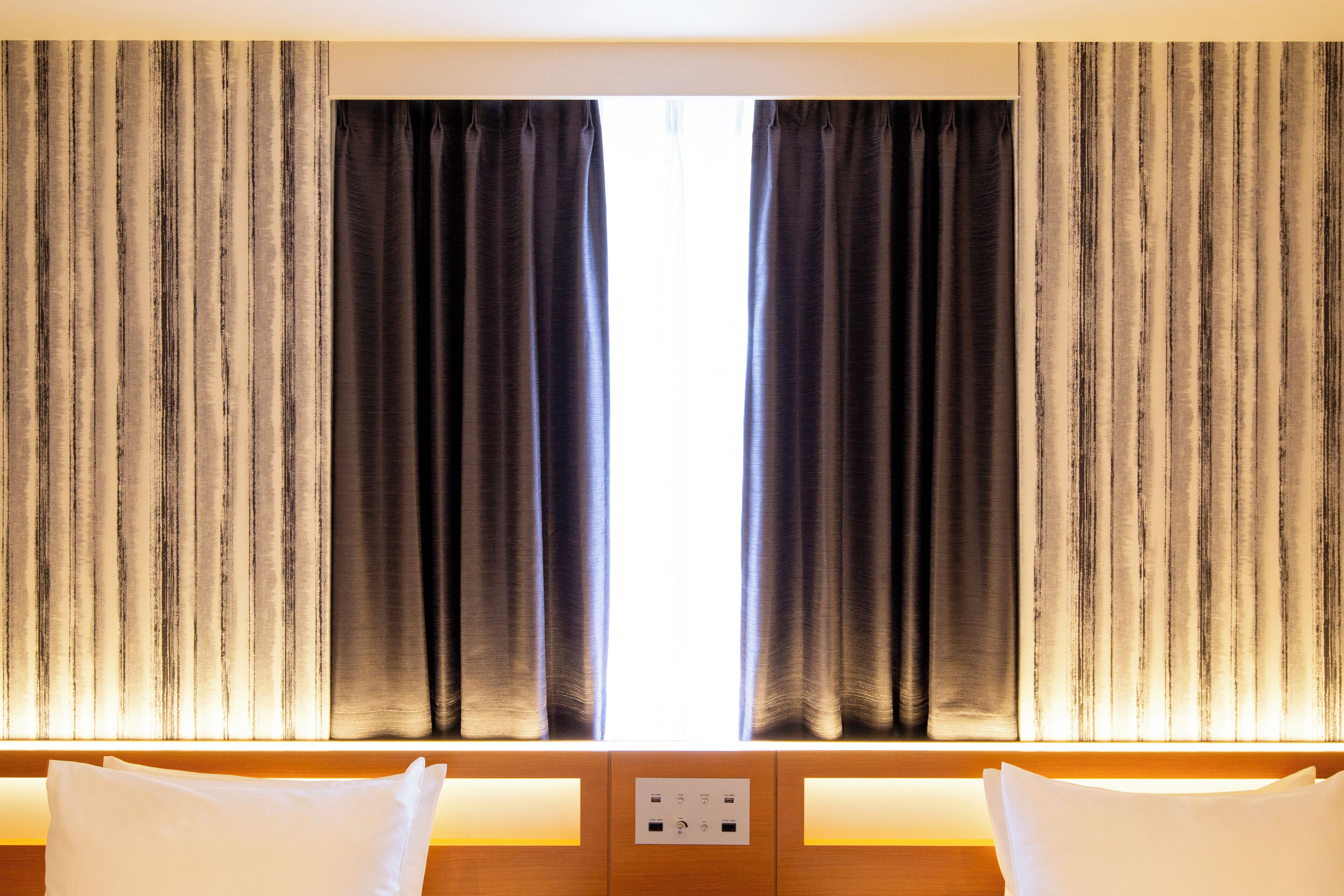 Una habitación de hotel con un ventanal grande, cortinas, una cama con almohadas y un panel de control.