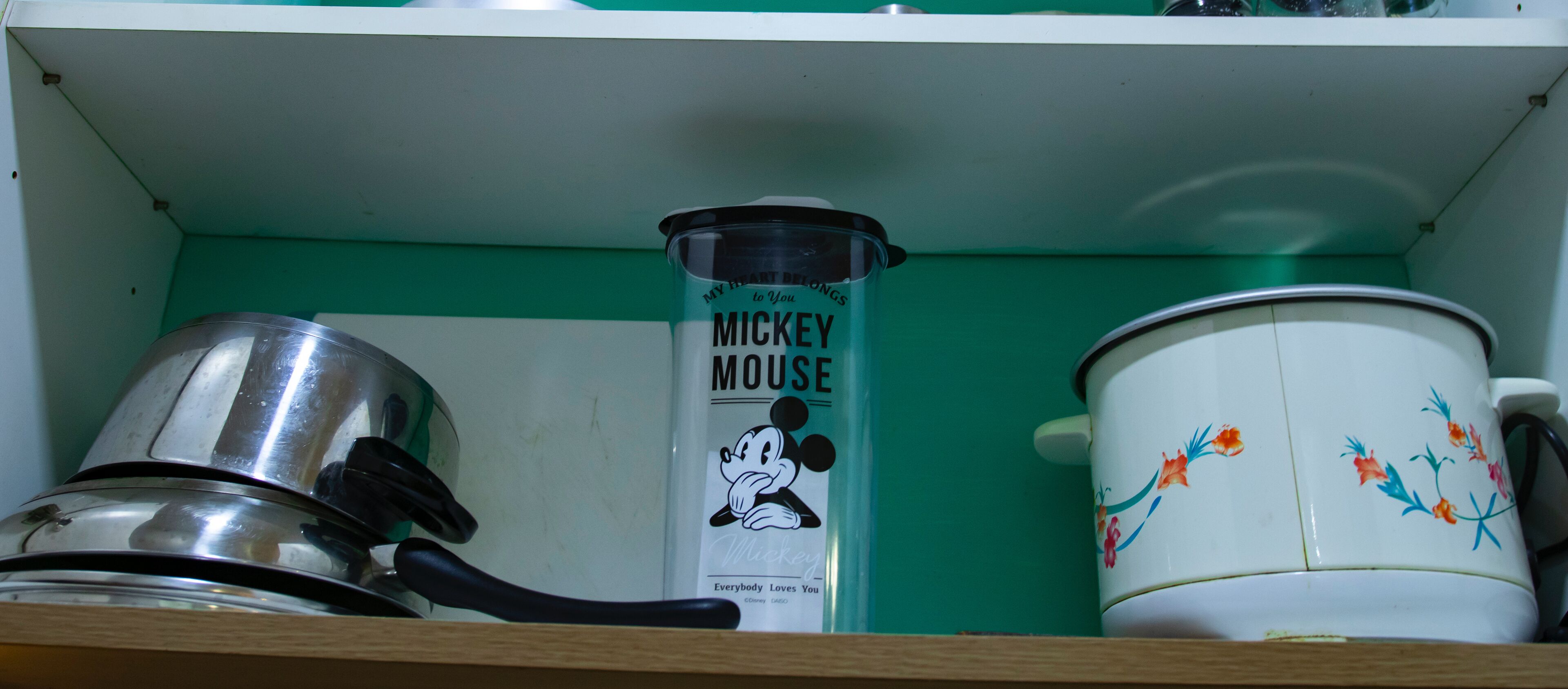 A kitchen shelf with a Mickey Mouse mug, a stainless steel pot, and a white ceramic pot with floral designs.