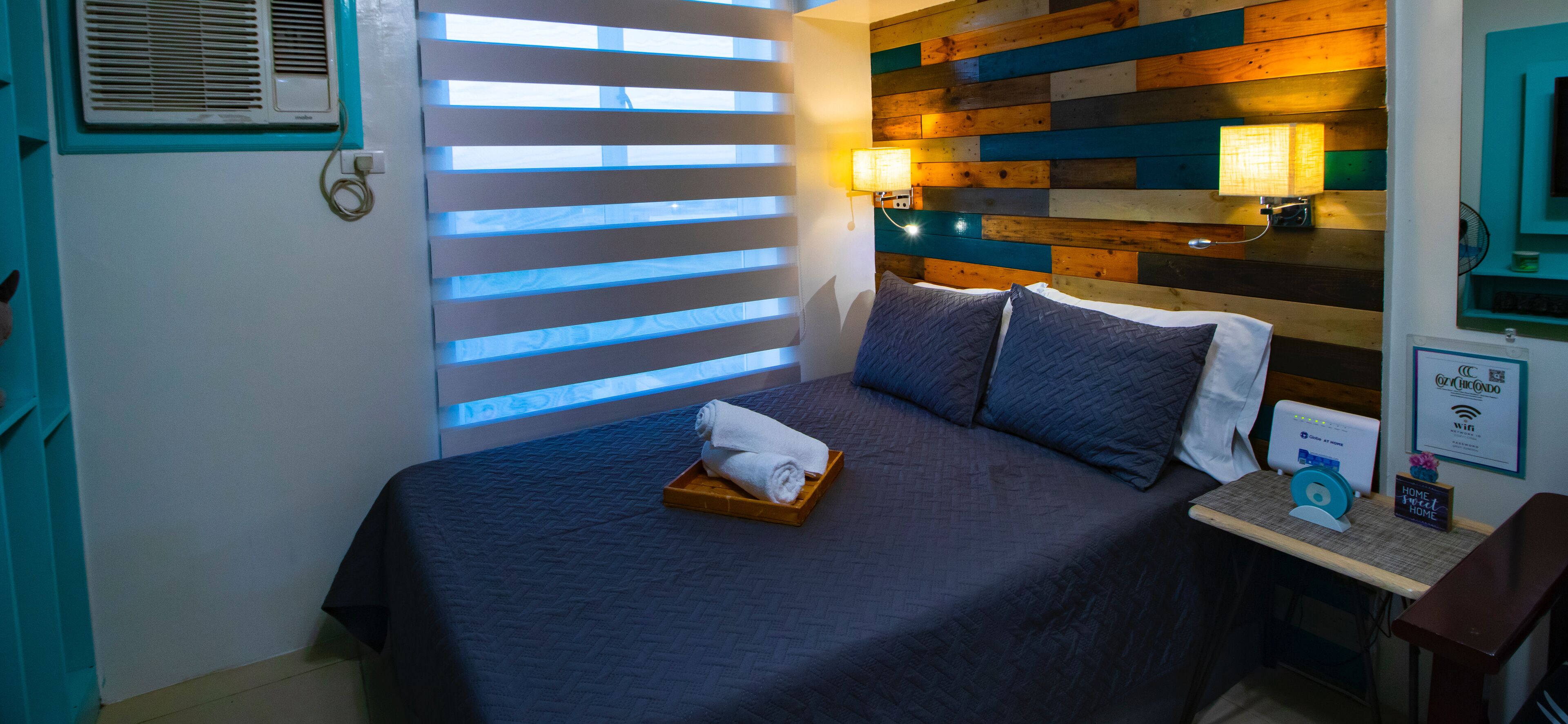 Een keurig opgemaakt bed met een dienblad handdoeken, een nachtkastje met een lamp, en een wandairconditioning.