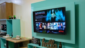 A teal-colored wall with a mounted TV displaying a movie selection screen. Below the TV, there are shelves with various items including a 'hello' sign, a vase, and a framed picture. - Avida Towers Sucat (Parañaque)