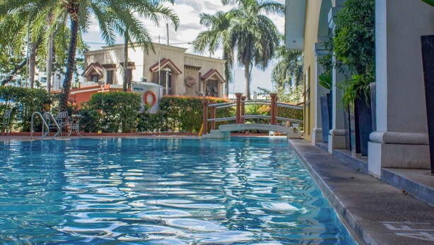 A swimming pool with clear blue water, surrounded by palm trees and a building with a light-colored facade and arched windows. - Avida Towers Sucat (Parañaque)
