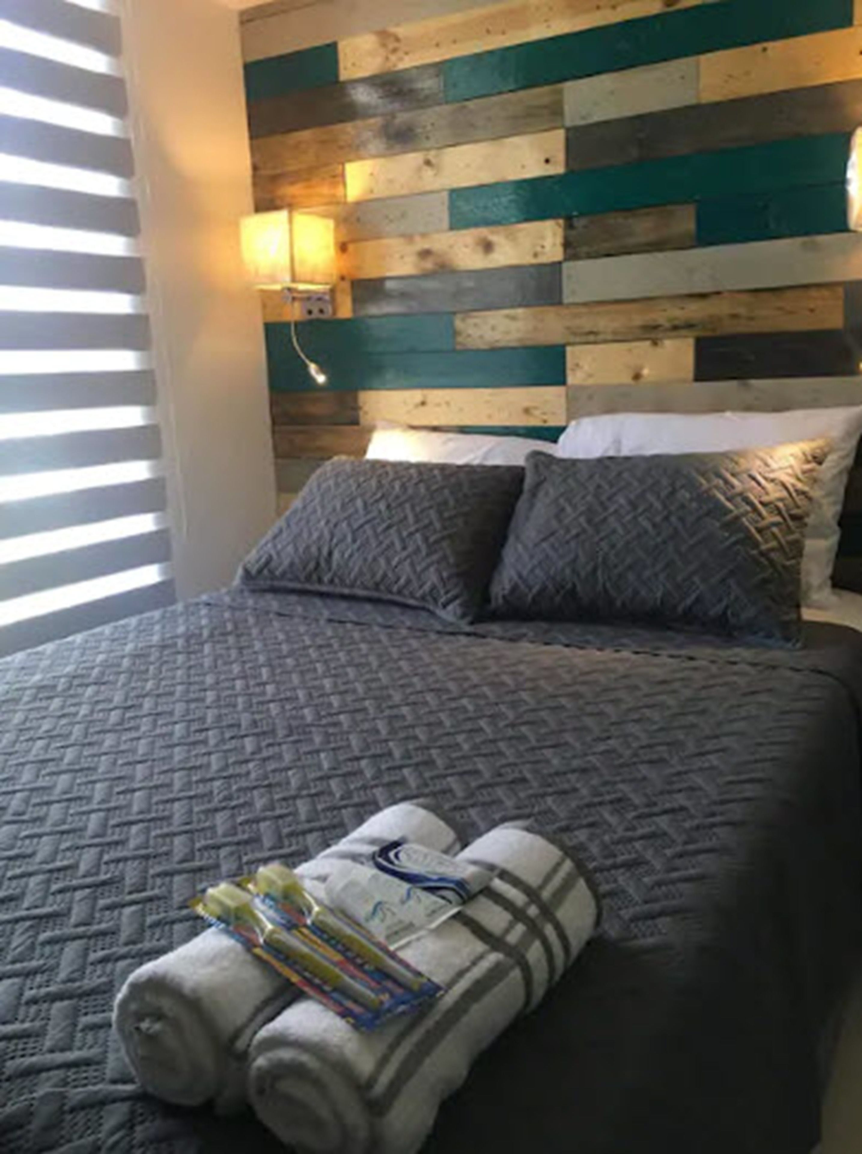 Una cama con colcha gris, dos almohadas grises y una toalla enrollada con libros encima.