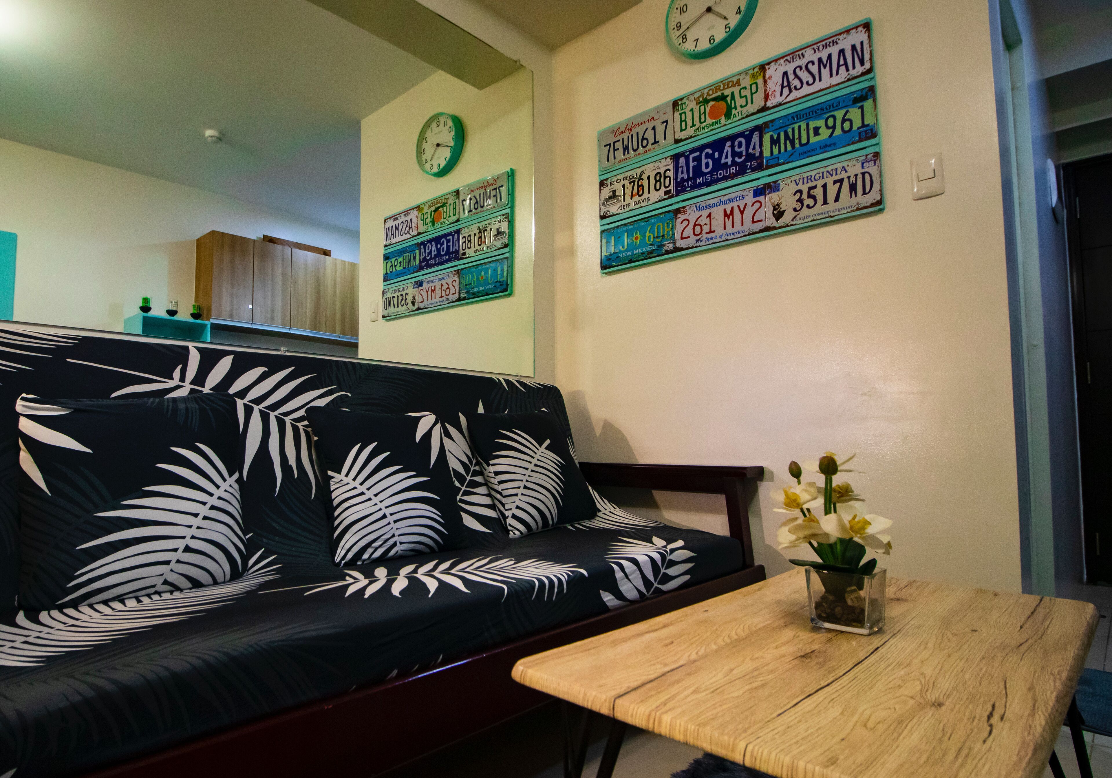 A living room with a black sofa with white leaf patterns, a wooden coffee table, and a wall with various license plates.