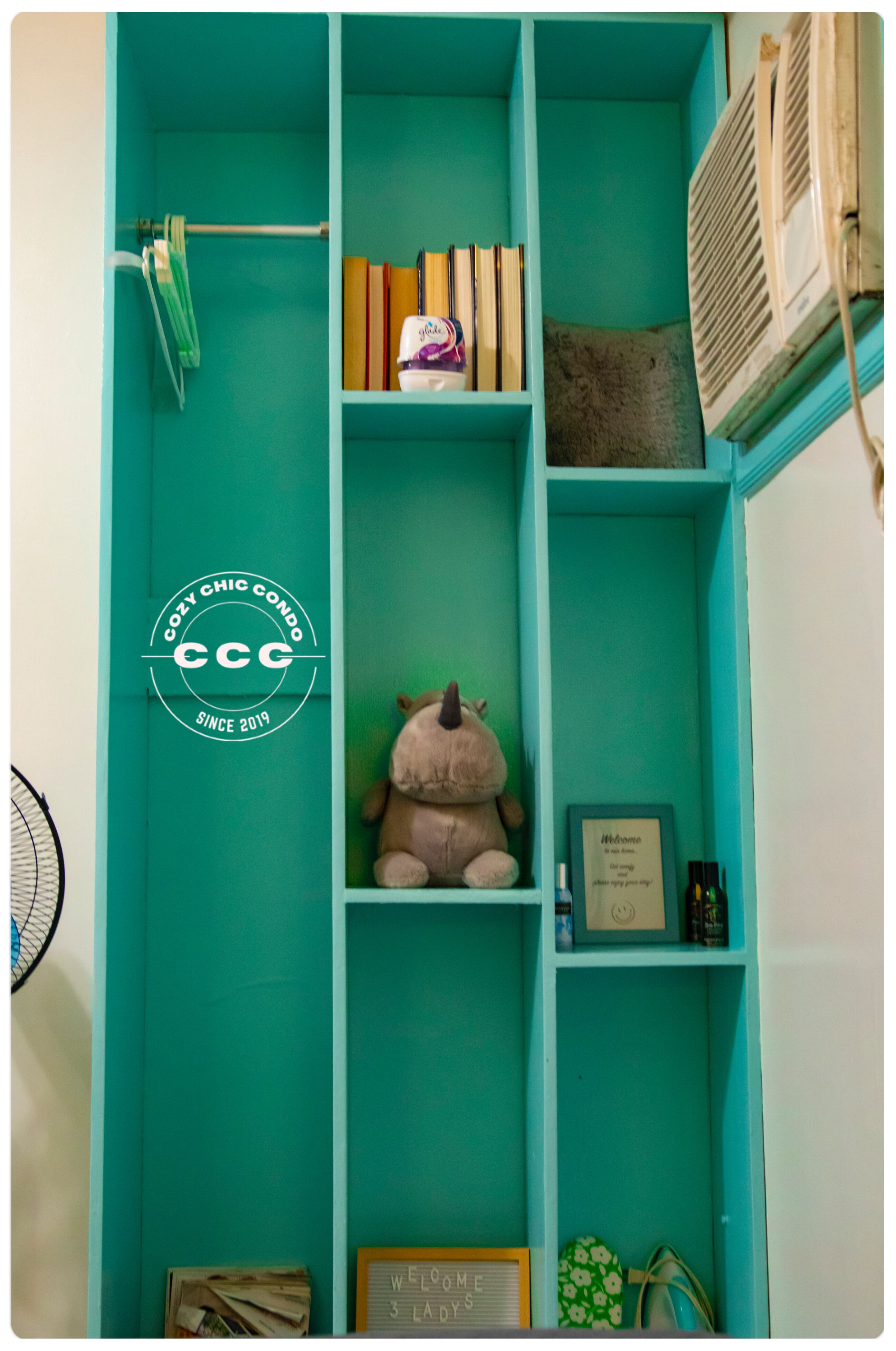 A teal-colored room with a bookshelf, a plush toy, a welcome sign, and a small framed picture.