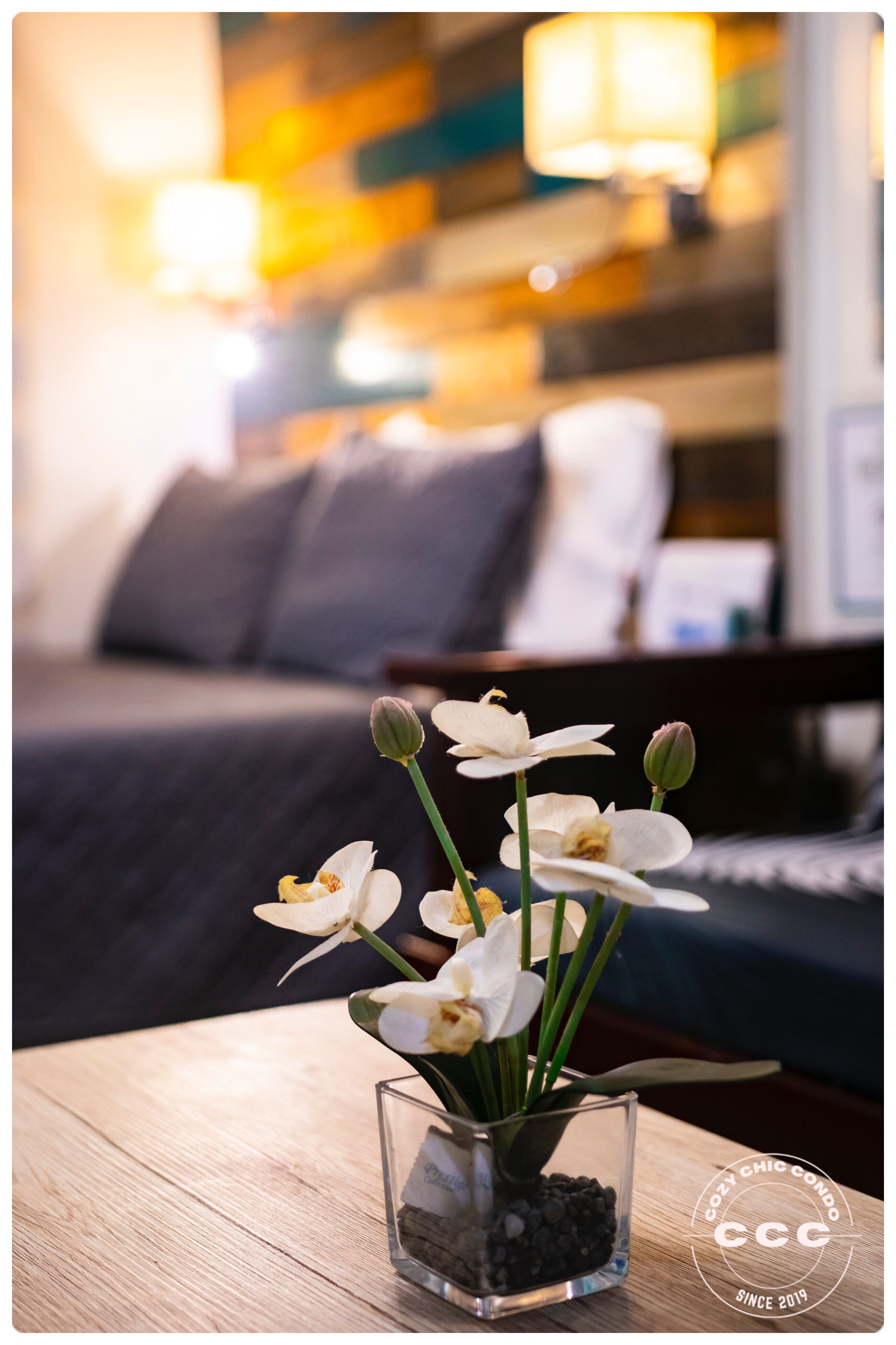 A glass vase with white orchids on a wooden table in a hotel room.