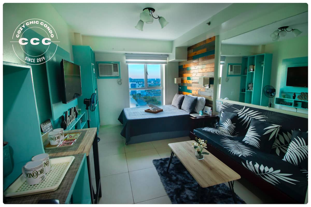A compact living space with a bed, sofa, and kitchenette. A large window offers a city view.