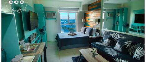 A compact living space with a bed, sofa, and kitchenette. A large window offers a city view.