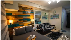 A bedroom with a wooden headboard, a bed with a dark comforter, a bedside table with a lamp, a black and white patterned sofa, and a small table with a vase. - Avida Towers Sucat (Parañaque)