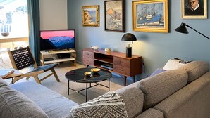 Una sala de estar con sofá, televisión, mesa de centro y lámpara. Hay cuadros en la pared y una ventana con cortinas.
