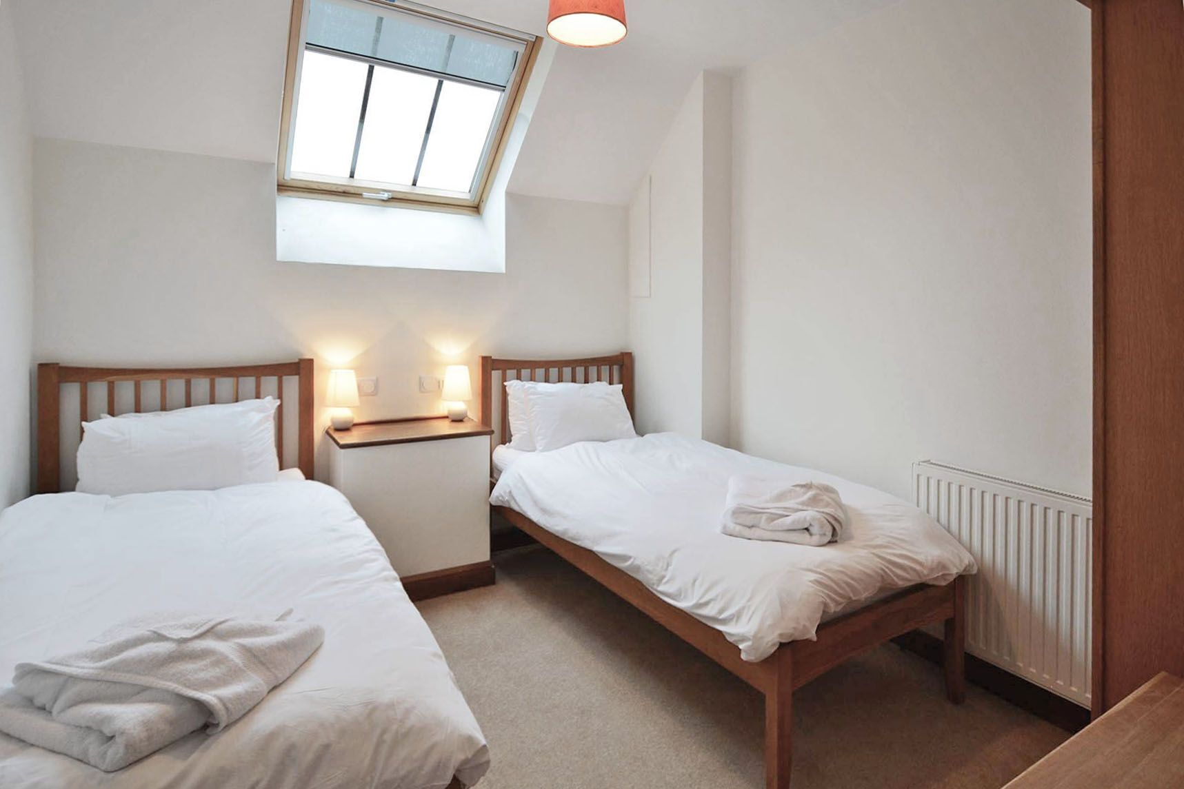 A bedroom with two single beds, a skylight, a bedside table with lamps, and a radiator.
