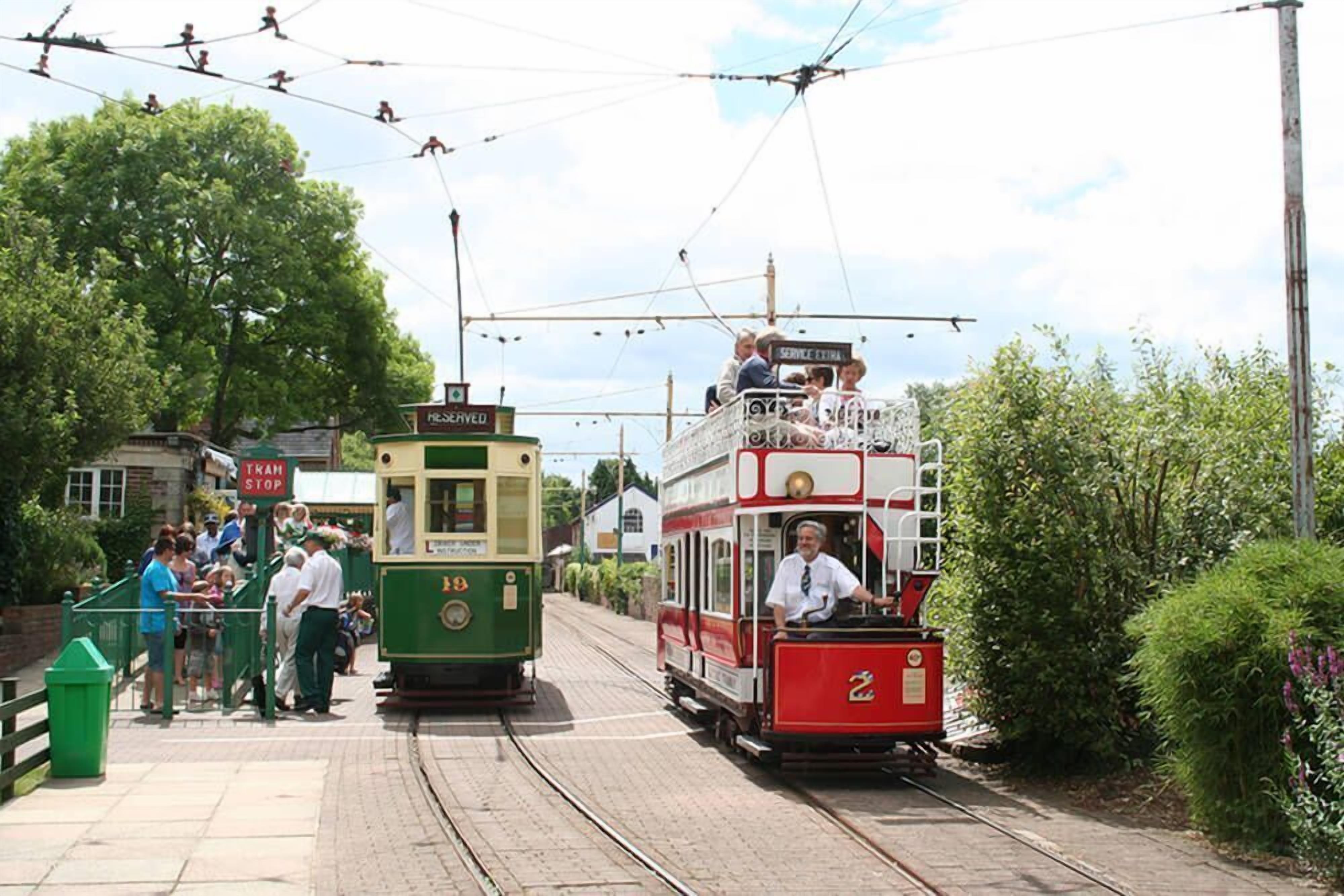 Une station de tram où l’on voit un tramway vert et un tramway rouge, des passagers montant et descendant.
