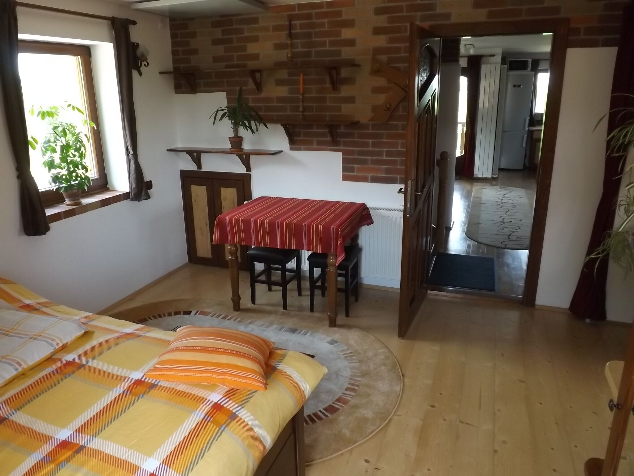 A bedroom with a bed, a table with chairs, a potted plant, and a view of a kitchen through an open door.