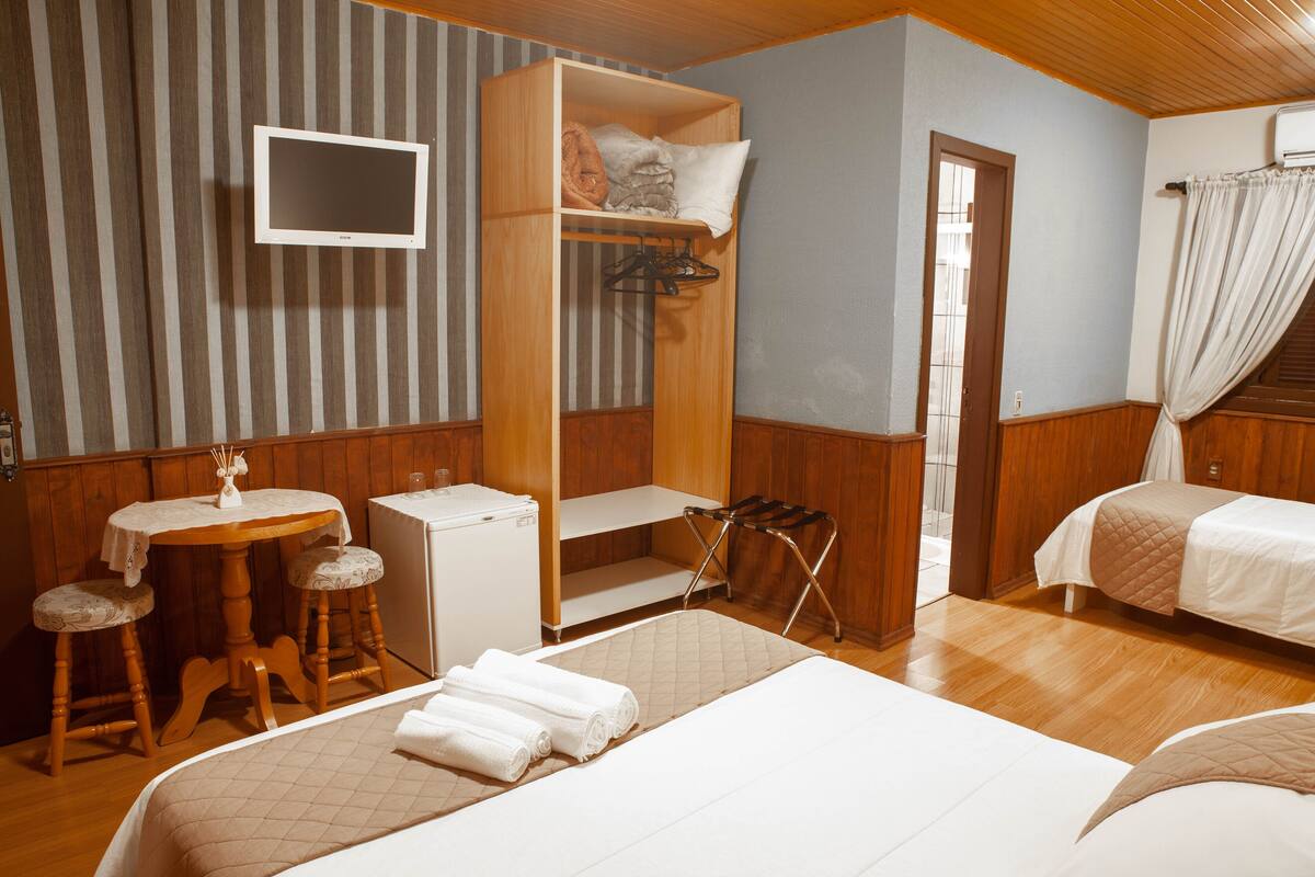A hotel room with a bed, a desk with a chair, a small table, a TV mounted on the wall, and a wardrobe.