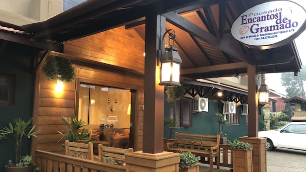 A cozy outdoor seating area with wooden benches and planters, a glass door entrance, and a sign for 'Pousada Encantos de Gramado'. - Pousada Encantos de Gramado (Gramado)