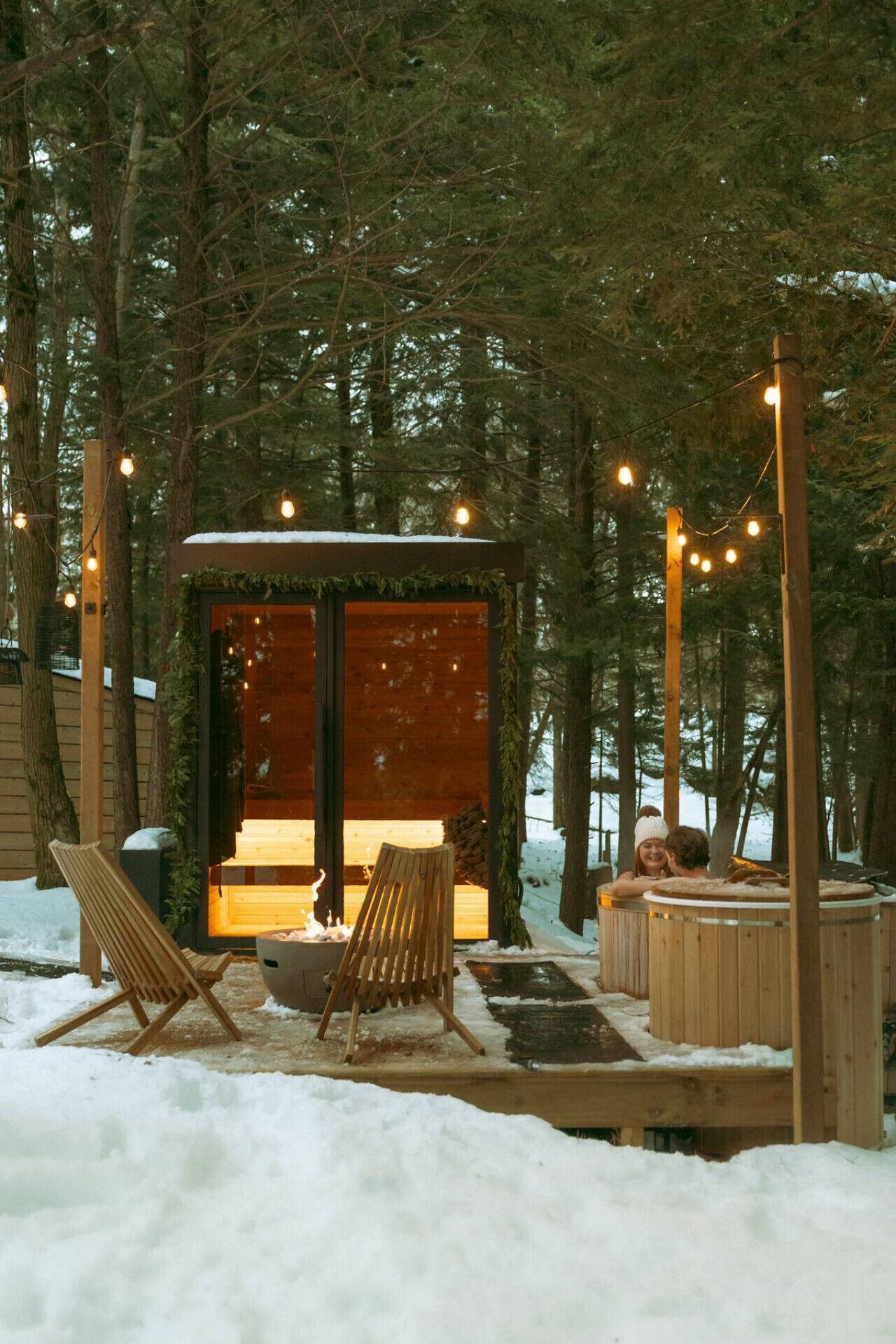 Une cabane en bois avec une porte en verre, entourée de neige, et dotée d’un espace extérieur aménagé avec des sièges et un foyer.