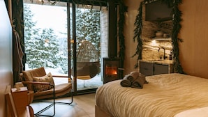 A cozy bedroom with a bed, a hanging chair, a fireplace, and a view of snow-covered trees.