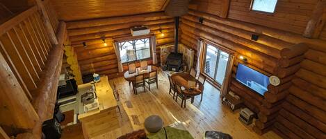 Interior de uma cabana de tronco com área de jantar, lareira, televisão e uma janela grande.