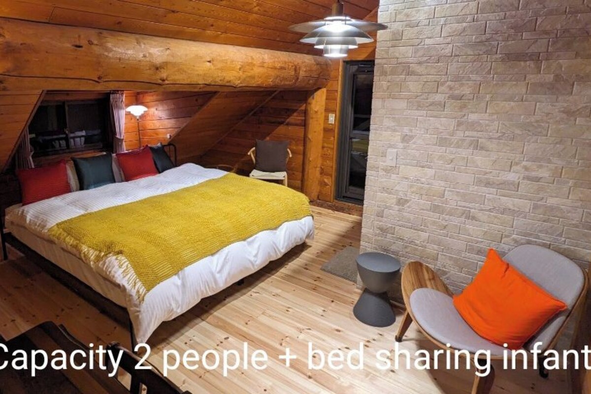 Une pièce confortable avec un lit, une chaise et une petite table. Les murs de la pièce sont en bois et le plafond est en pente.