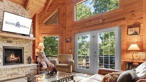 A cozy log cabin interior with a fireplace, leather sofa, and a large window offering a view of the outdoors.