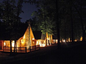 A log cabin-style building with a prominent triangular roof, surrounded by trees and illuminated by warm lighting. - Genuine Log Cabin-Near Big Cedar! Private Hot Tub & Grill! Spacious Deck & Car Charger (Ridgedale)
