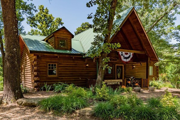 A rustic log cabin with a green metal roof, surrounded by lush greenery and trees.