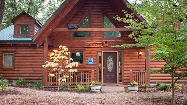 A log cabin with a front porch, a sign reading "Branson Deer Lodge," and a small tree in the front yard.