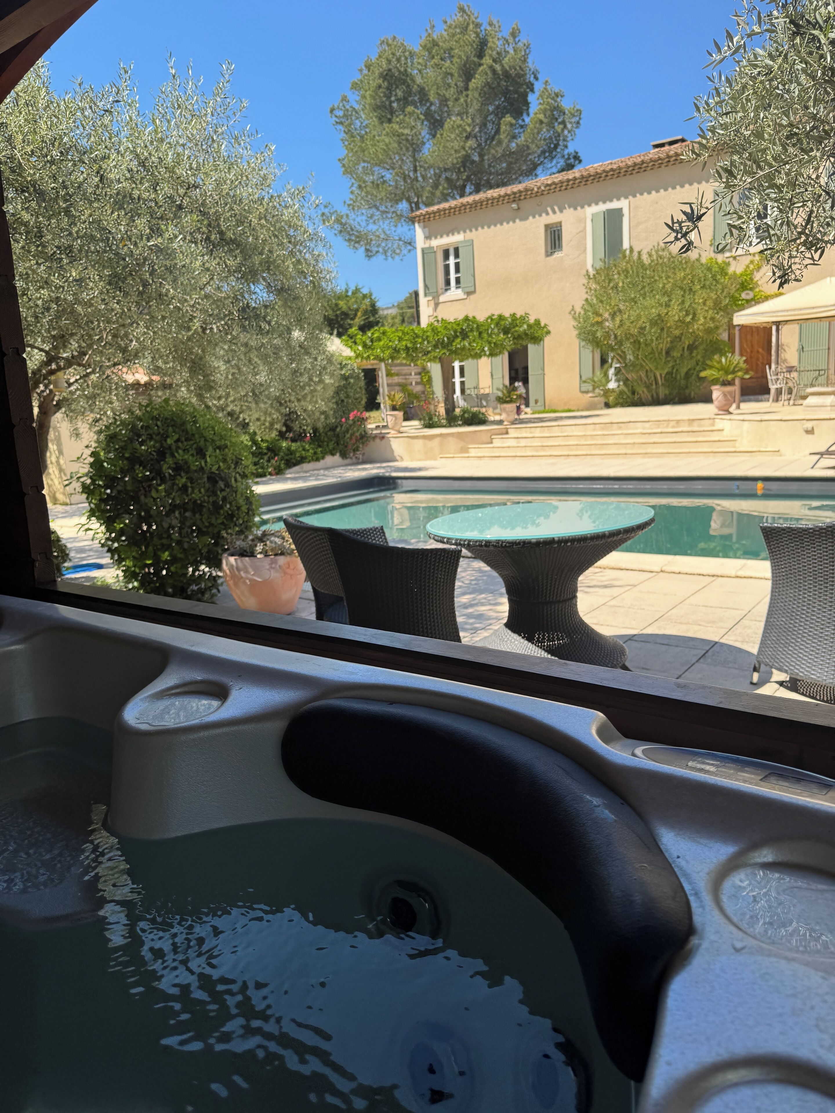 A hot tub with a view of a pool and outdoor furniture, surrounded by greenery and a building in the background.