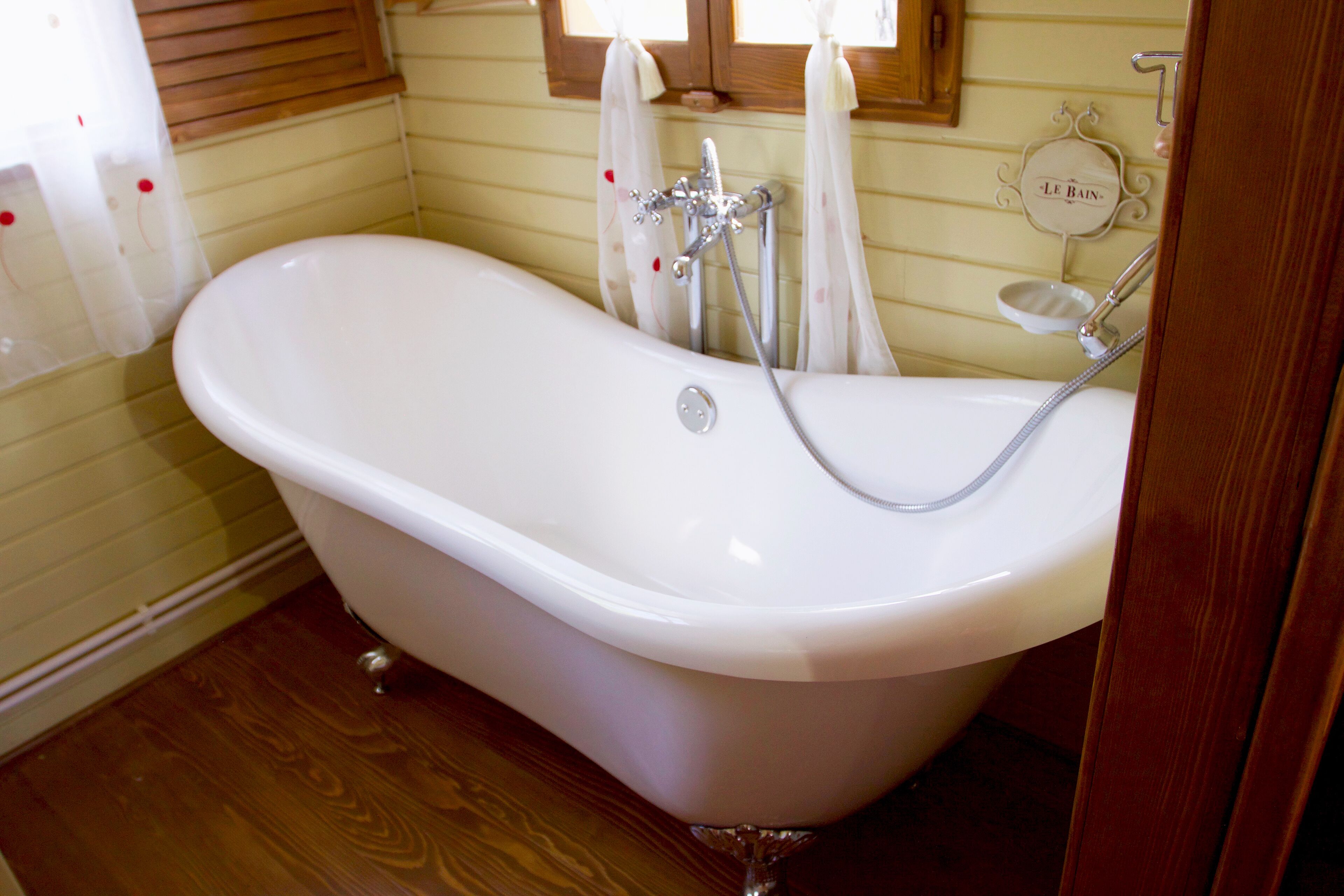 Una bañera blanca independiente con ducha y grifo, suelo de madera y una ventana con cortinas.