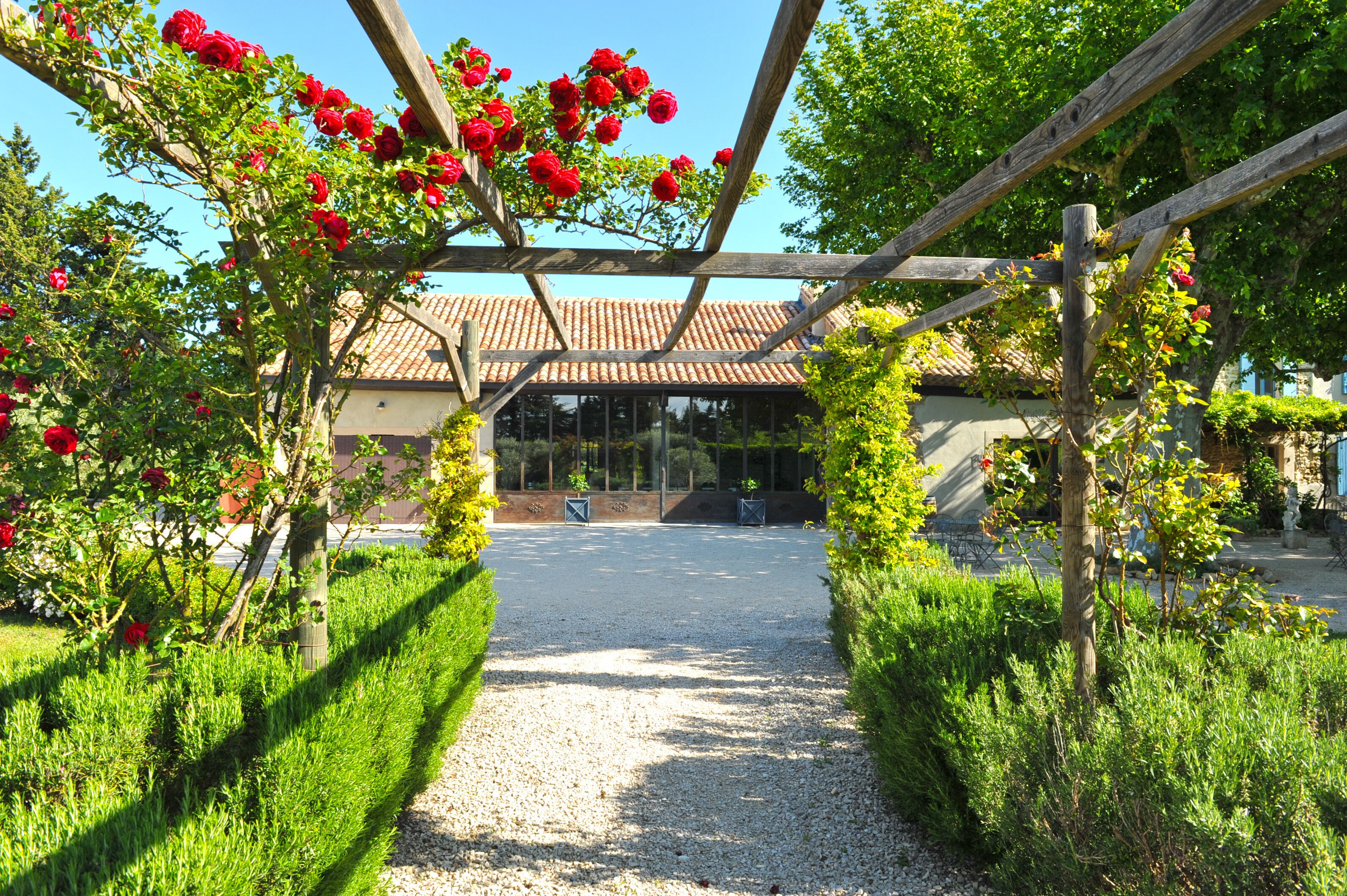 Un chemin de gravier mène à un bâtiment doté d’un toit en tuiles, entouré d’une végétation luxuriante et de fleurs rouges.