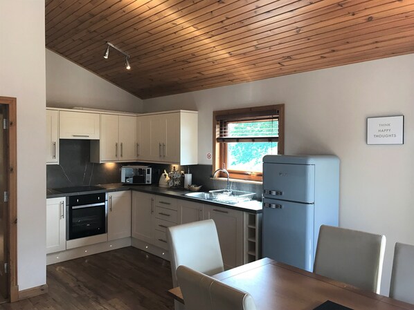 A modern kitchen with white cabinets, a microwave, a kettle, and a refrigerator. There is a dining table with chairs and a window with blinds.