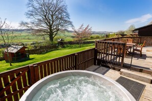 A hot tub on a wooden deck with a view of a green landscape and a small wooden structure in the background.