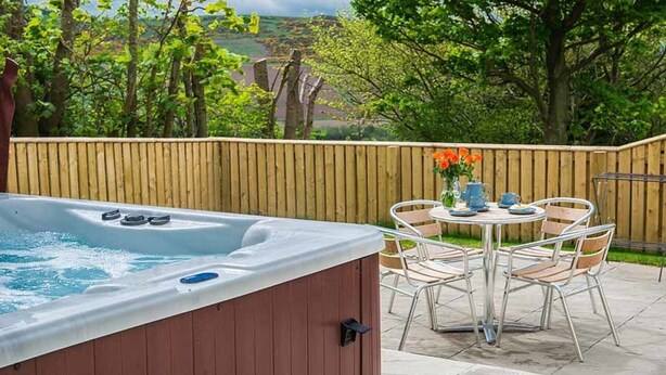 Un bain à remous avec une terrasse en bois, entouré d’une clôture en bois, et une table avec des chaises et un vase de fleurs.
