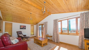 Ein geräumiges Wohnzimmer mit einem roten Ledersofa, einem Holztisch und einem Fernseher. Große Fenster mit Vorhängen bieten einen Blick ins Freie.